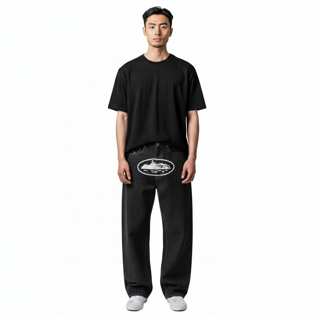 The Hype Company baggy jeans washed black denim with oval island graphic on front. Model wearing black t-shirt and white sneakers.