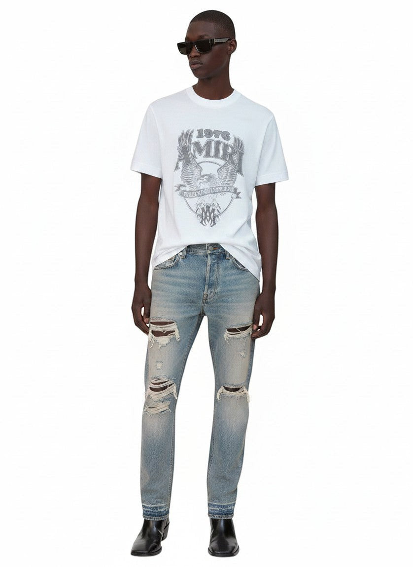 Modelo vistiendo camiseta blanca The Hype Company AMIRI 1976 Hollywood con estampado gráfico y vaqueros desgastados
